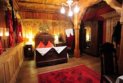 Blick in das historisch gestaltete Zimmer. 
Mittelalterliches Bett aus massiven Holz und Holzschnitzereien und roter Bettwäsche. Passend zum Bett historische Beistelltische, Kleiderschrank und Stühle. Wände, Decke und Boden mit Holz verkleidet. Balken sin