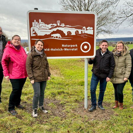 Gruppe von sechs Personen steht neben einer neuen braunen Willkommenstafel des Naturparks Eichsfeld-Hainich-Werratal. Die Tafel zeigt stilisierte Symbole des Naturparks, darunter Gebäude, Brücken und einen Bären. Im Hintergrund ist eine offene Wiesenlands